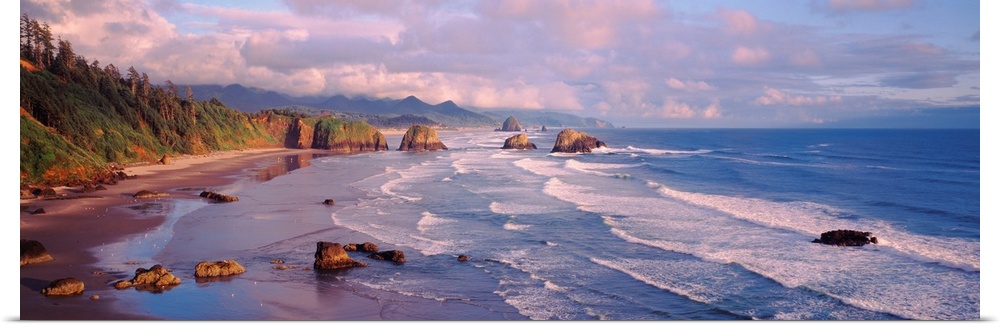 Panoramic photograph of waves slowly crashing against the rocks and sand of Cannon Beach in Oregon.  The mountains in the ...