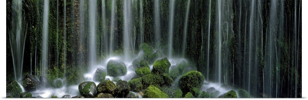 Panoramic photograph displays a gathering of large rocks covered in moss as the waterfall above crashes into and splashes ...