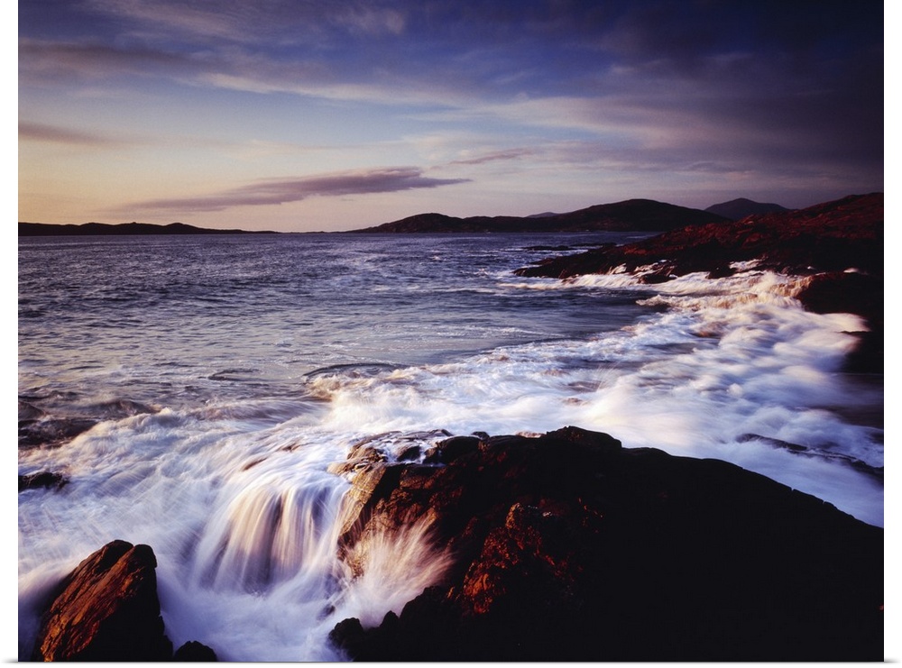 Horizontal photograph of water crashing into the rocky shoreline of Outer Hebrides as the sun sets over Scotland, a mounta...