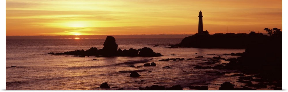 A lighthouse and the cliffs surrounding it are silhouetted by the sunset over the Pacific ocean.
