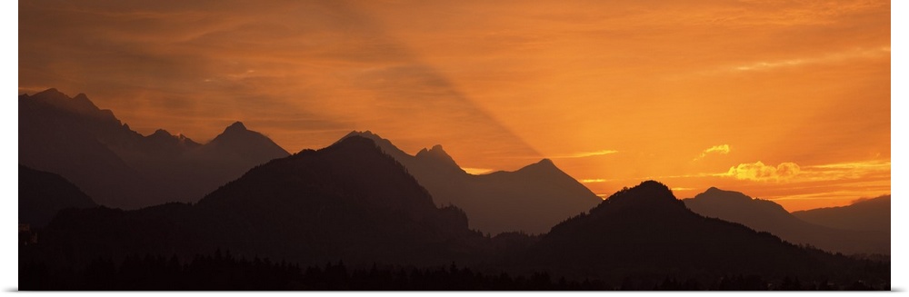 Wide angle photograph of the sun setting behind the silhouetted European Alps, beneath a golden sky, in Bavaria, Germany.