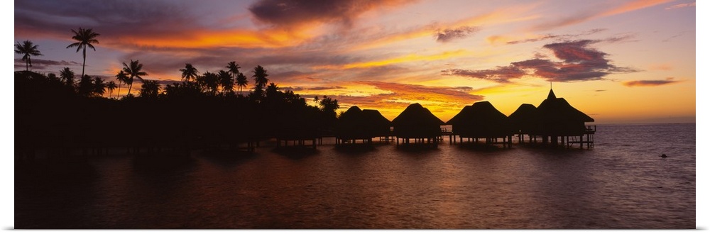 Long and narrow photo on canvas of palm trees and houses on stilts over the ocean silhouetted against a bright sunset.