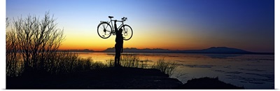 Silhouetted cyclist holding bicycle over head, river's edge, sunset, Alaska