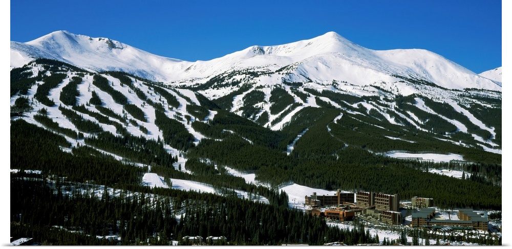 Ski lodges are shown at the bottom of snowy, forested mountain range in this large photograph.