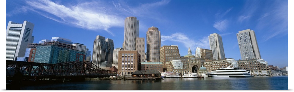 The Boston skyline is photographed in panoramic view from the waterfront during a bright sunny day.