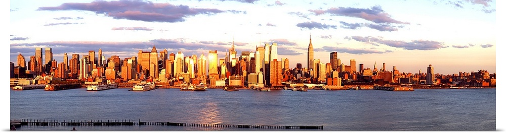 Panoramic photograph of the distant Manhattan skyline at the waters edge, with golden skyscrapers as the sun sets.