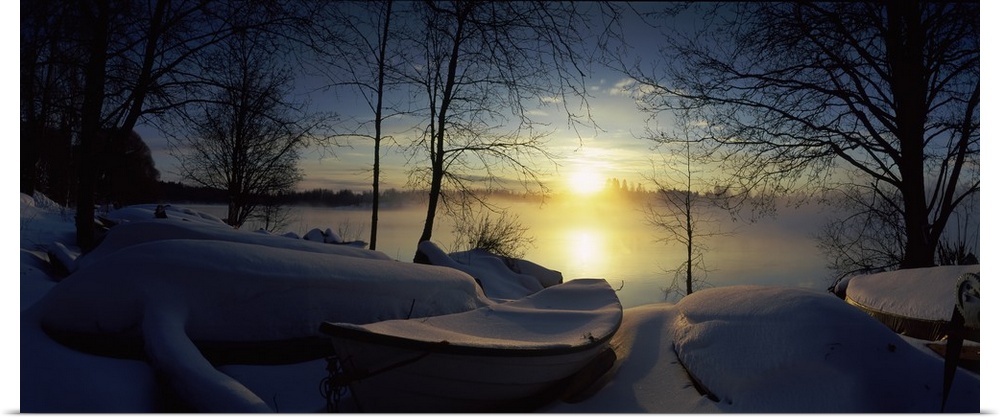 Panoramic photograph displays a group of rowboats blanketed in fresh powder sitting near the shore of a river in Europe.  ...