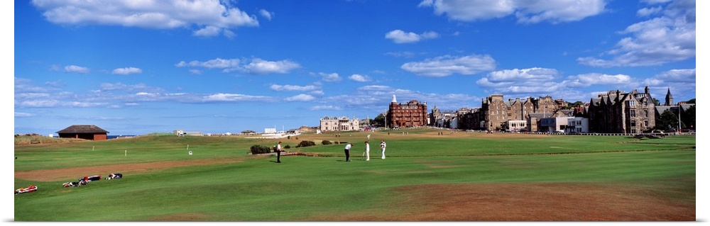A group of people tee off on a golf course in the UK. Buildings on the course line the right side of the panoramic piece.