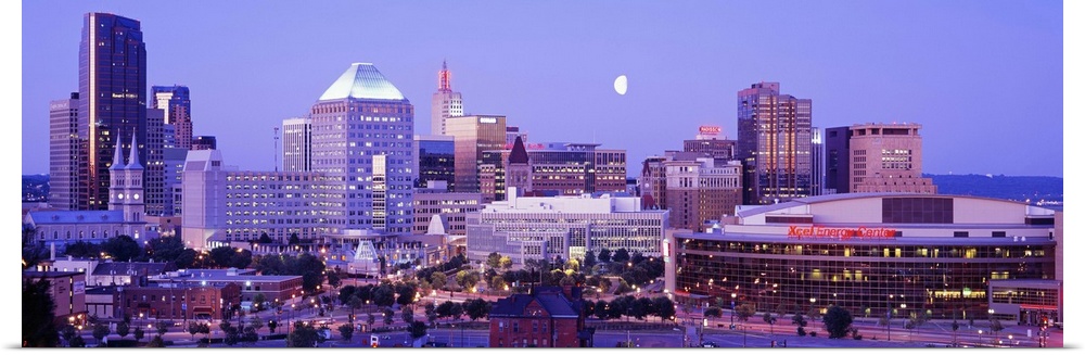 Panoramic photograph of skyline lit up at dusk with moon in the sky.