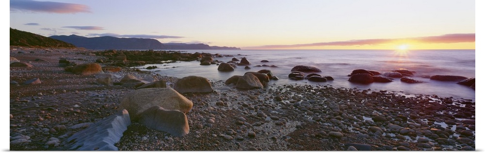 Panoramic photograph of rock and pebble filled shoreline with sun setting in the distance.