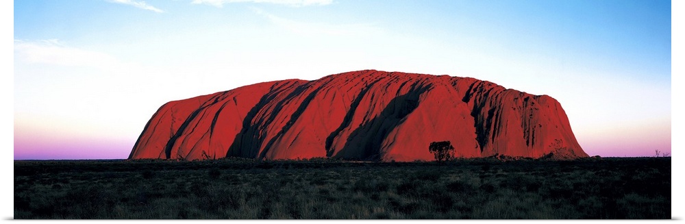 The Ayers Rock is photographed in wide angle view during a sunset which lines the horizon with warm colors.