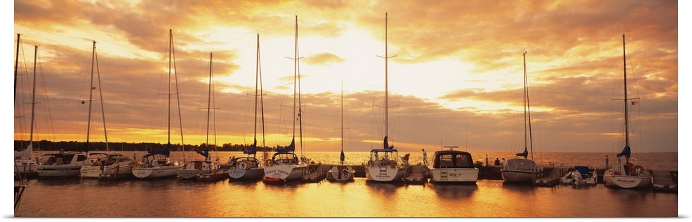 Horizontal panorama of a row of sailboats with their sails down, docked at a port on a lake at sundown in New England.
