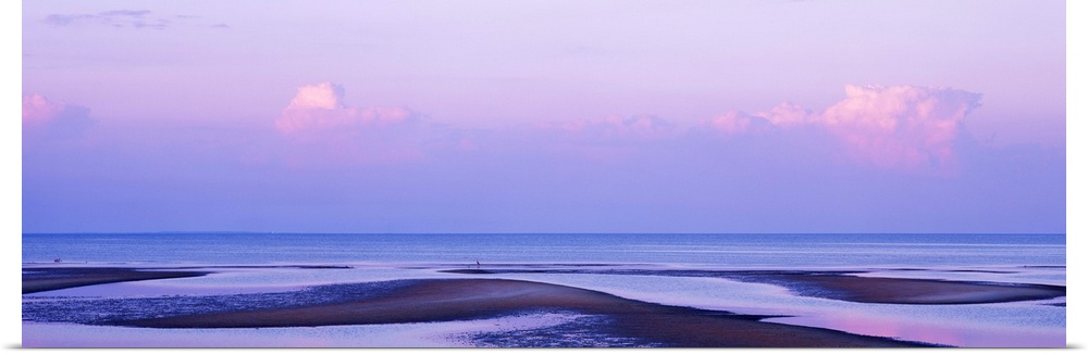 An elongated view of the ocean with large water puddles on the beach and the sky has pink tones from the sunset.