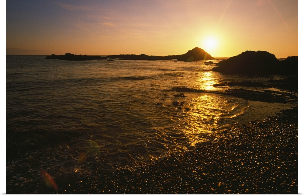 Large, landscape photograph of the sun setting over large rocks in the water, near the shore of the Pacific Coast in Oregon.