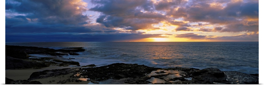 A panoramic photograph of the sun setting beyond the horizon as viewed from a rocky tropical beach.