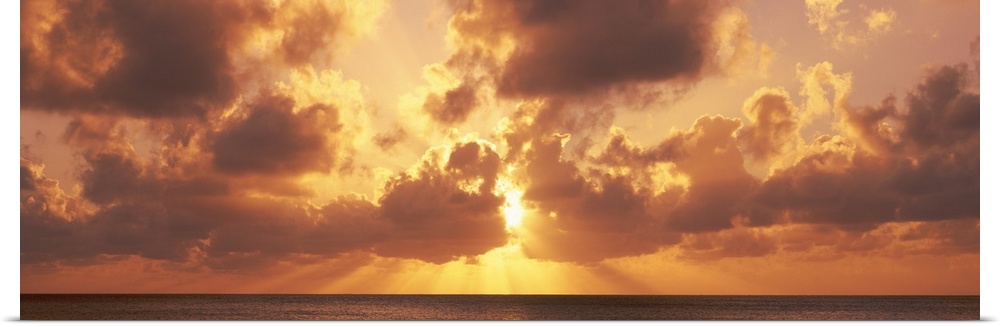 Large panoramic photograph taken of a sunset behind the clouds with its rays bursting through and a vast ocean beneath it.
