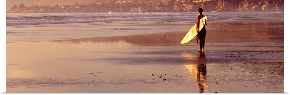 Panoramic photograph of a single surfer walking along the shore with his surfboard at Pismo Beach, California.