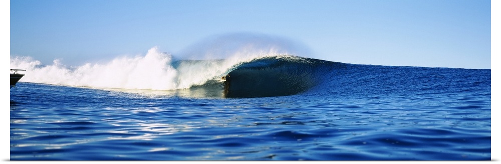 Large, horizontal photograph of a distant surfer riding a large wave, in the blue waters of Tahiti, French Polynesia.