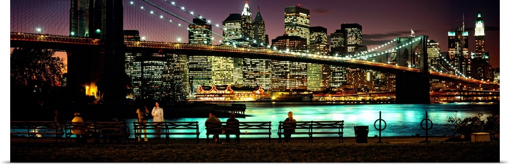 Manhattan lights reflecting in the East River under the Brooklyn bridge while people sit on benches watching the view.