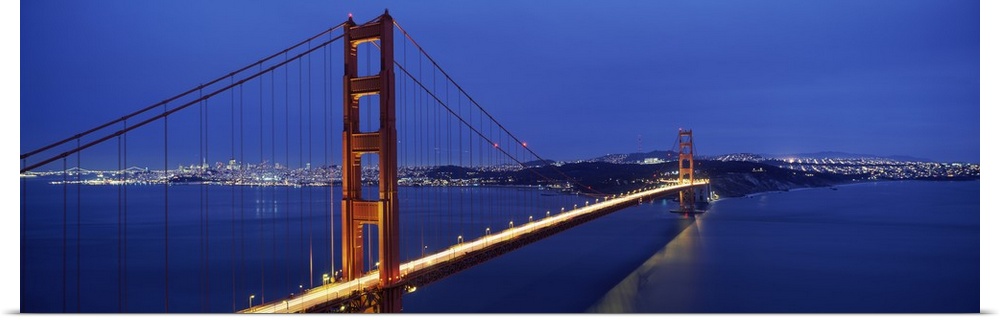 Panoramic photo of a bridge spanning the bay, its famous red towers and cables standing out against blue twilight sky.