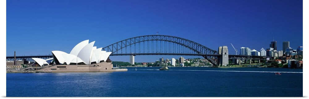 This is a panoramic photograph of the harbor with the suspension bridge, opera house, and city skyline in the background.