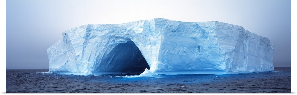 This panoramic photograph is of an immense iceberg that has a tunnel going through the middle of it.