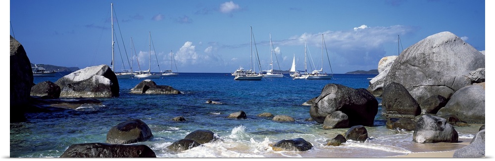 Big rocks strewn on a sandy beach being splashed by the tide with several sailboats in the distance.