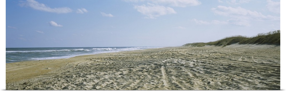 A panoramic photograph of a sandy beach indented with footsteps and tracks down to the ocean water.