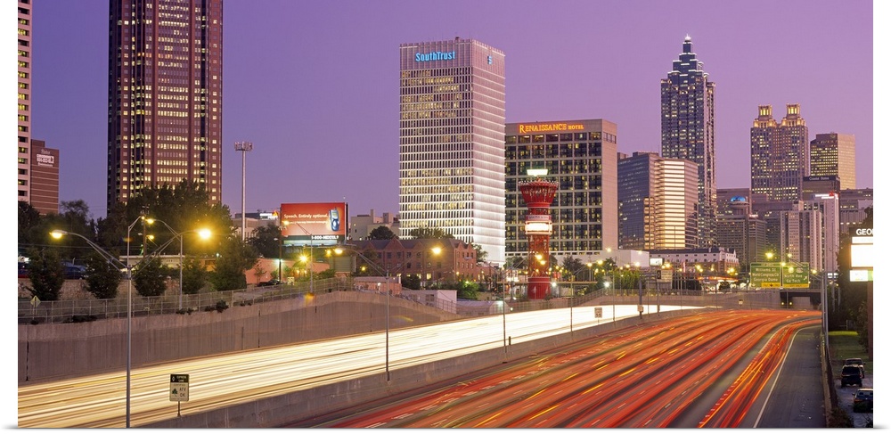 Panoramic photograph of highway filled with light trails lined by lit up buildings at sunset.