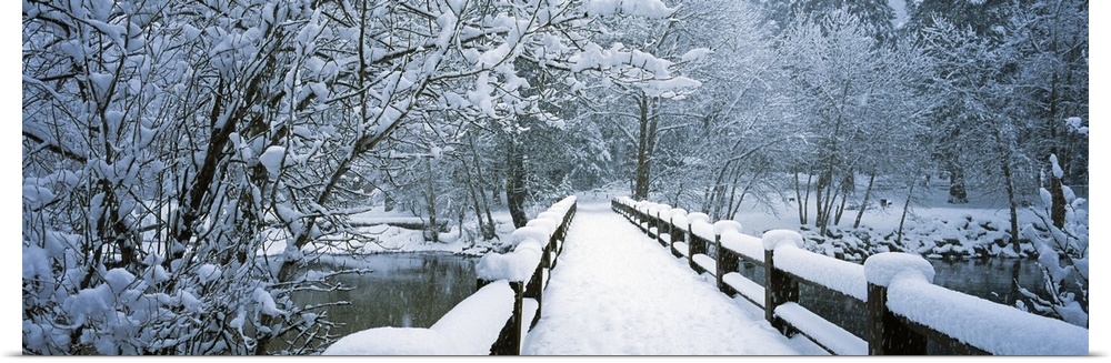 Large panoramic piece of a small bridge that is covered with inches of snow and surrounded by snow covered trees.