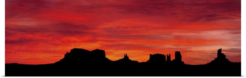 Panoramic photograph of silhouettes of huge rock formations in the desert at sunset under a cloudy sky.