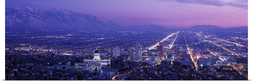 High-angle panoramic photograph of city lit up at dusk with snow covered mountains in the distance under a cloudy sky.