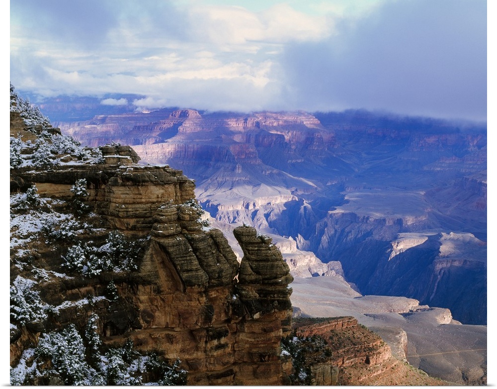 Thick clouds hover over the Grand Canyon that is photographed from a distance behind a snowy cliff.