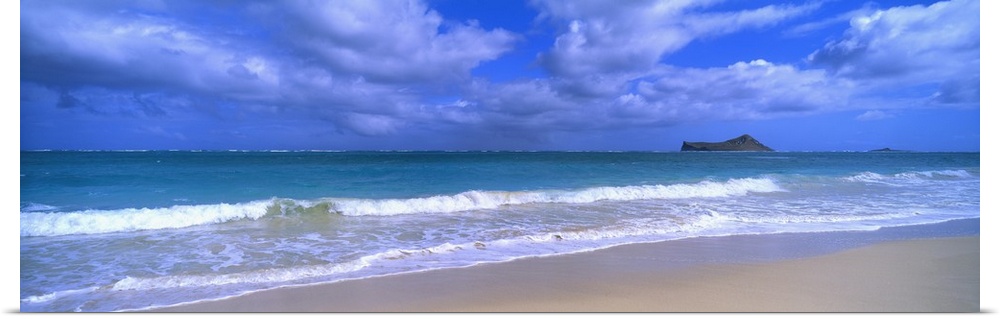 Panoramic photograph of waves breaking on the beach during a cloudy day with island in the distance .