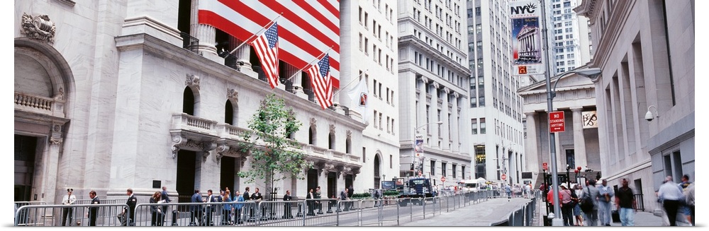Large panoramic photograph of wall street in NYC with people walking on the sidewalk and a large American flag to the left...