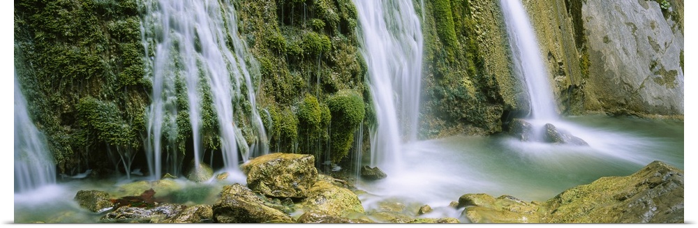 Panoramic photograph taken of several waterfalls cascading over foliage and crashing onto rocks.