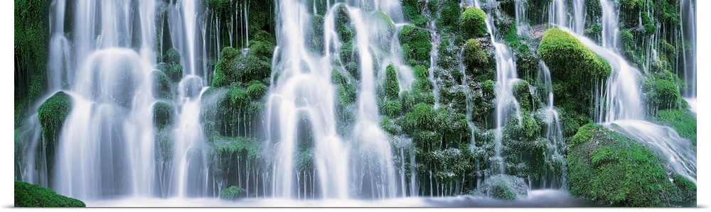 A panoramic picture taken of a massive waterfall that flows over vast green foliage.