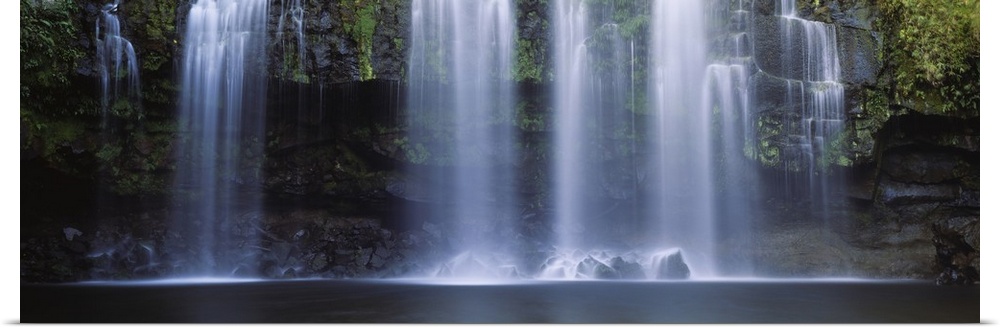 Multiple cascades fall over a moss covered rock face into a pool in this panoramic photograph of a tropical landscape.