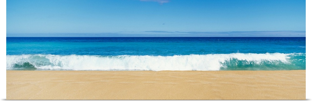 This panoramic photograph captures a swell breaking on the sandy shore of a tropical beach on a clear and sunny day.