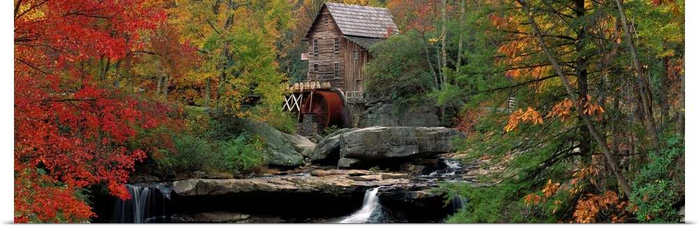 An old water mill sits at the top of flat boulders and a water fall in the center of this panoramic canvas depicting an au...