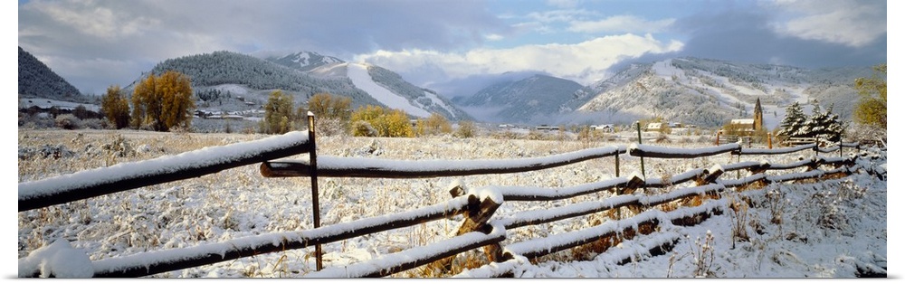 Panoramic photo of a snow covered landscape with a thin wooden fence running through it in the foreground and rolling moun...