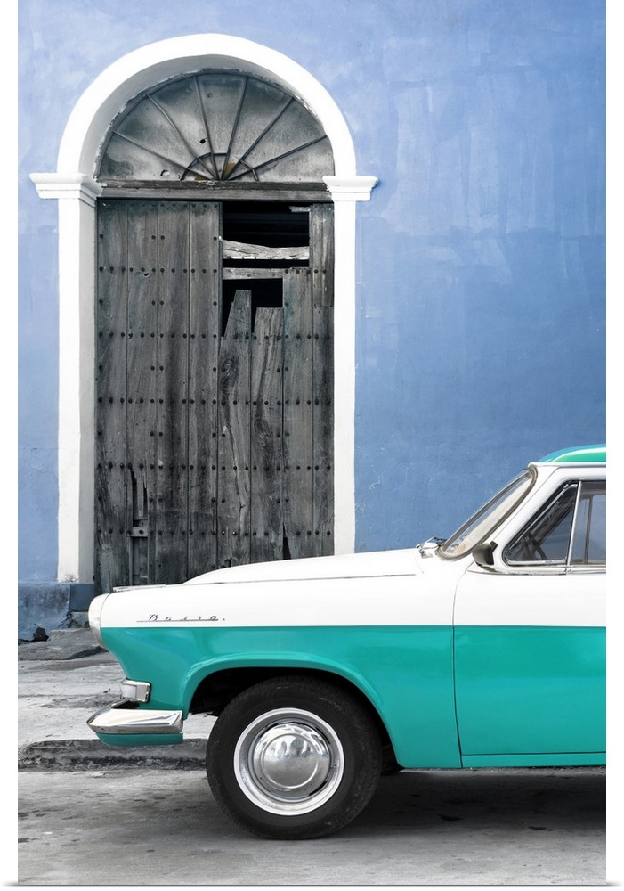 Photograph of the front of a vintage turquoise and white car outside of a blue building with a broken wooden door.