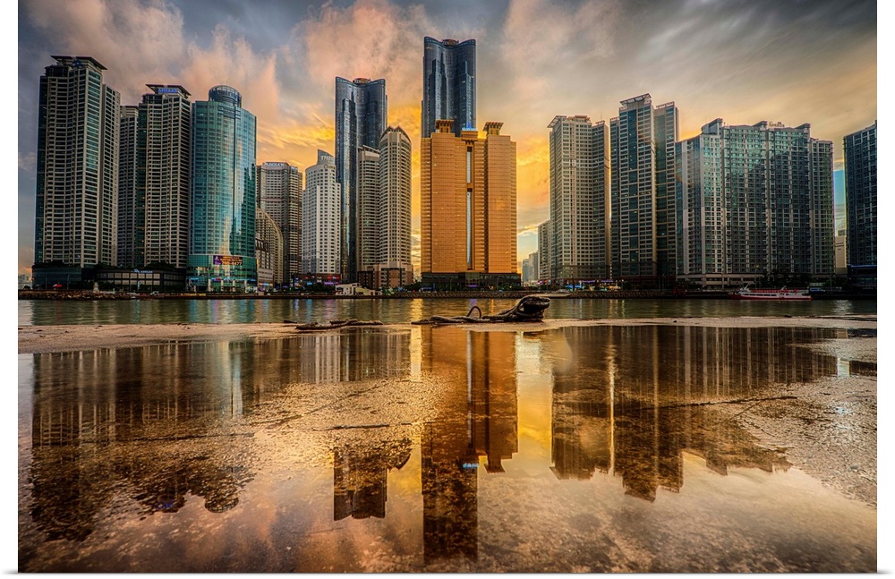 Dramatic photograph of a city skyline with immense skyscraper reflected in a puddle in the foreground of the image.