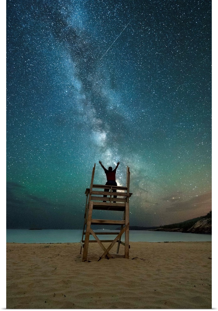 A photograph of a person on a lifeguard tower under a blanket of star in the night sky.