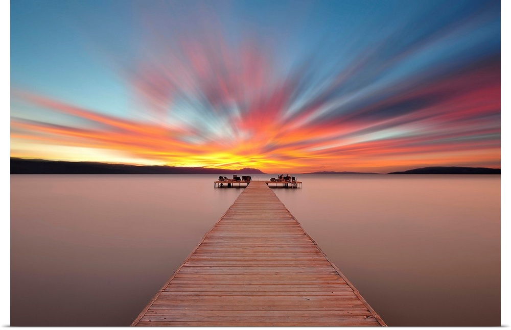 A wooden pier stretching out onto the water under a brightly colored sunrise sky.