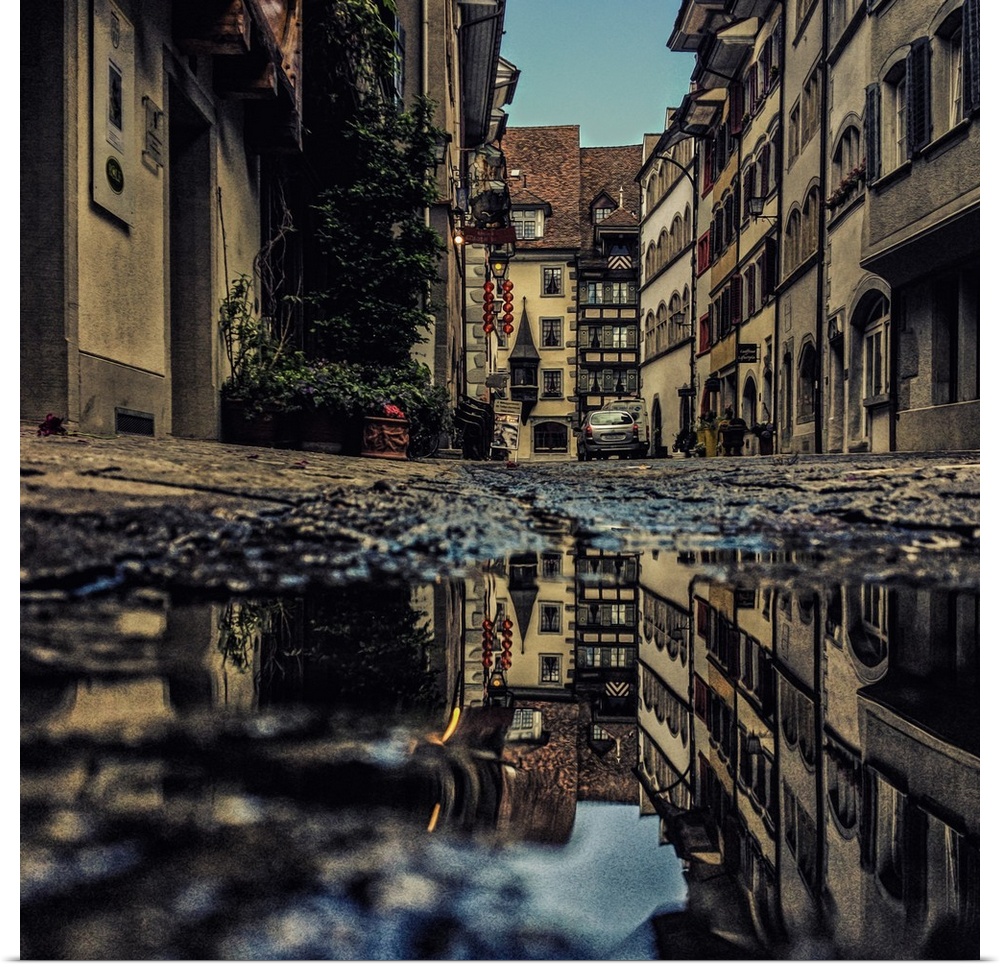 Photograph of a puddle reflecting surrounding buildings and atmosphere.