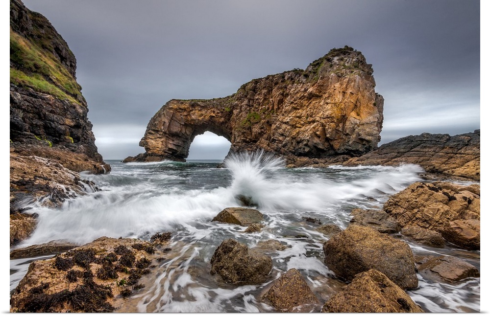 Oceanwater splashing onto the rocky shores in Ireland, with a natural bridge formation in the distance.