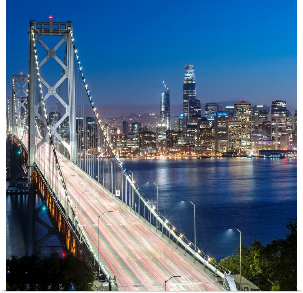Square photograph of the Bay Bridge at dusk with downtown San Francisco lit up in the background.