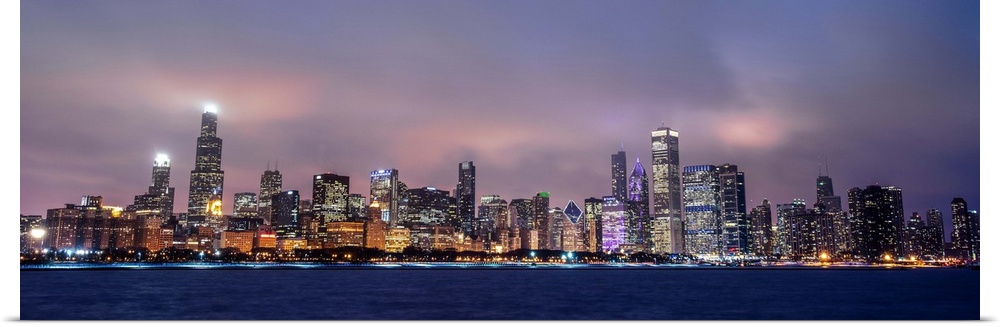 Panoramic view of the Chicago city skyline in the early evening, with city lights reflecting off the clouds above.