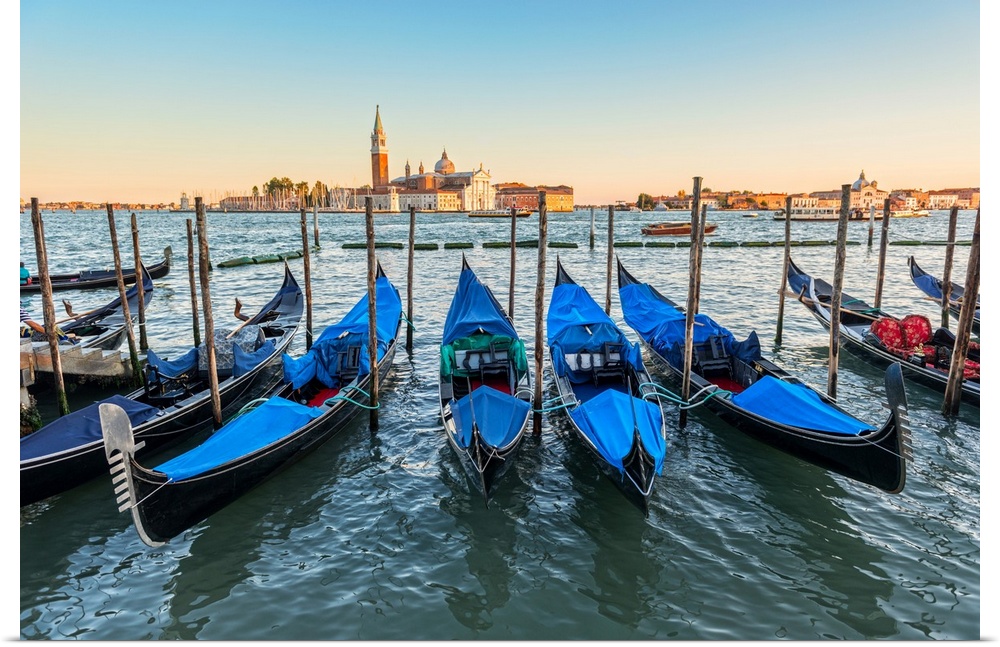 Photograph of a row of docked gondolas with St. Mark's Square (Piazza San Marco) in the background at golden hour.